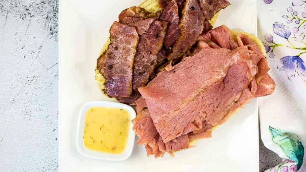 Slices of corned beef and pastrami on bread, served with a small dish of mustard sauce on a white plate, next to a floral napkin.