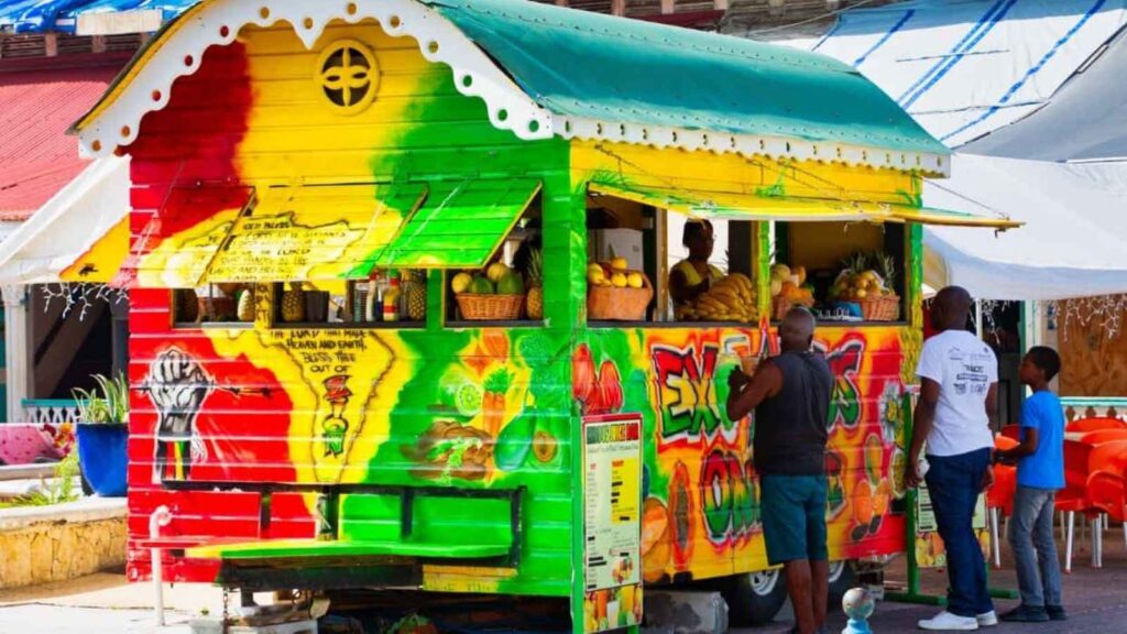 A colorful food truck painted in red, yellow, and green serves fruit and drinks to customers standing outside on a sunny day, offering a taste of restaurants abroad right on the street.