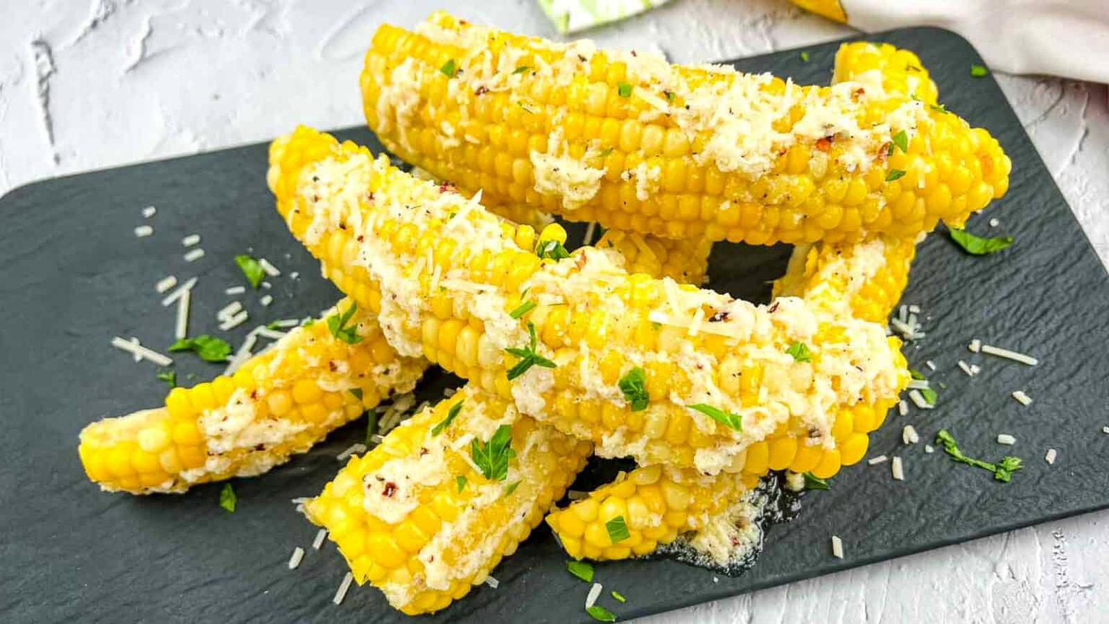 Several pieces of Parmesan Garlic Corn Ribs, topped with grated cheese and chopped herbs, are arranged on a black slate serving board.