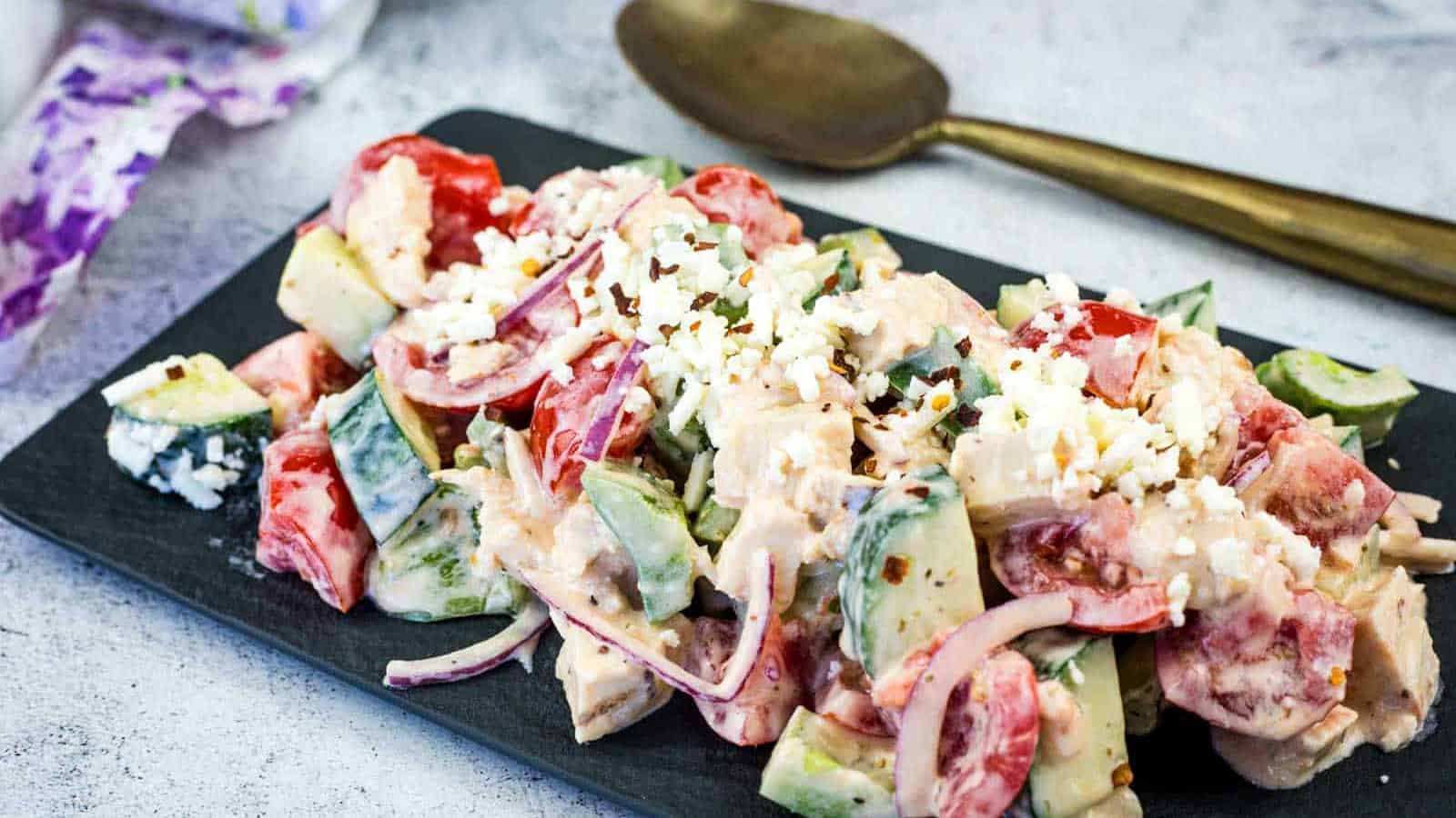 A plate of Greek Salad with Chicken sits on a dark serving tray next to a spoon.