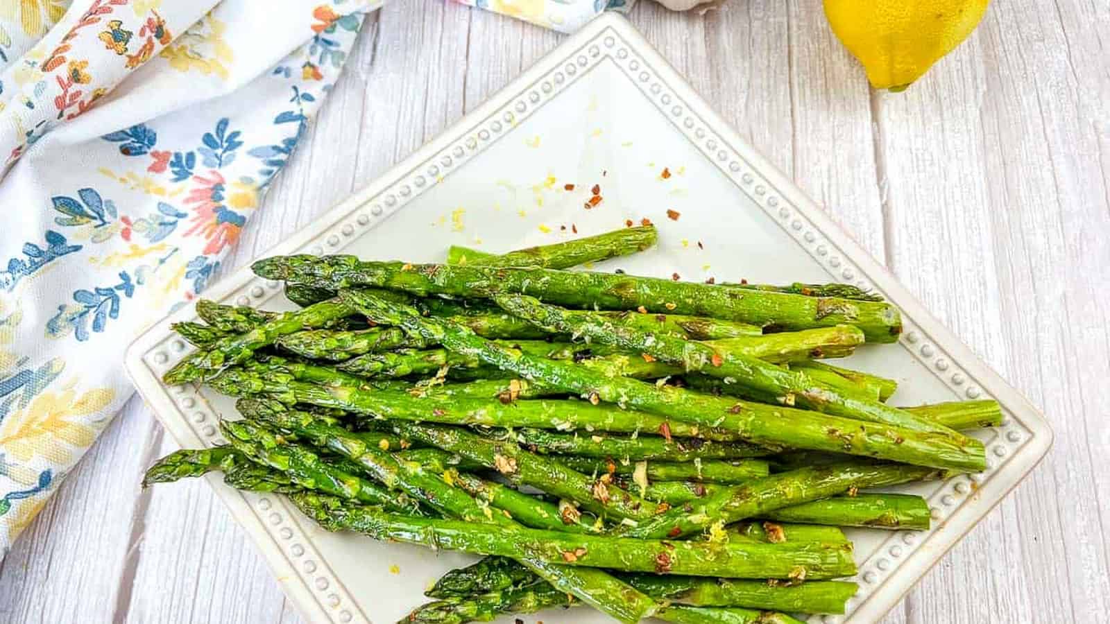 A square white plate with roasted asparagus spears, garnished with seasoning, sits on a light wooden table next to a garlic bulb, lemon, and a floral napkin.
