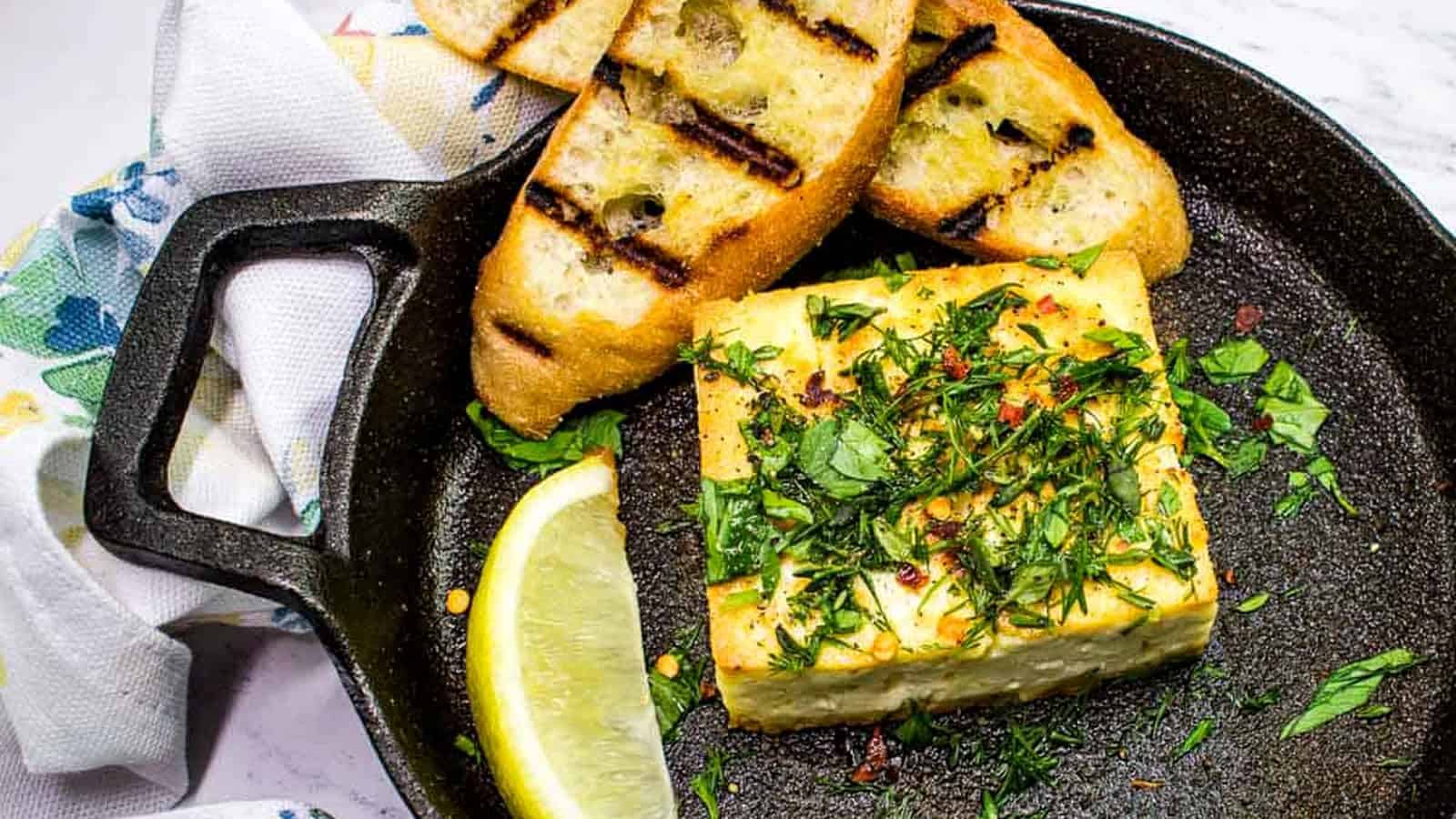 Grilled Feta with Herbs in a black serving dish with toasted bread and a lemon wedge.