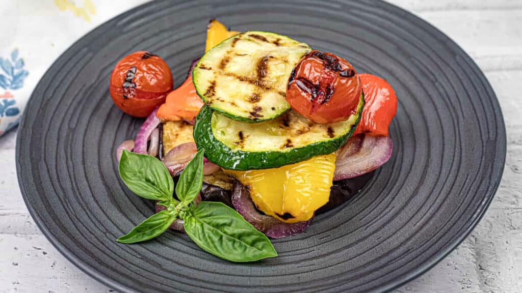 A plate of grilled vegetables including zucchini, tomato, bell pepper, and red onion, garnished with fresh basil leaves on a dark round plate.