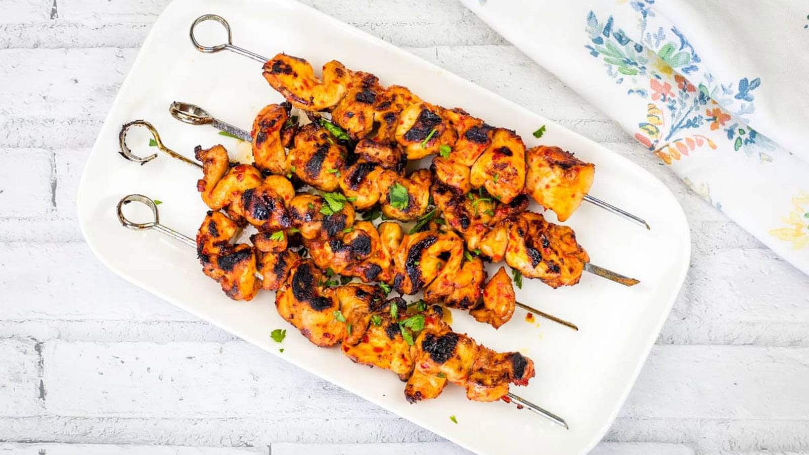 A rectangular white plate with four Sweet & Spicy Grilled Harissa-Honey Chicken Skewers garnished with chopped herbs.
