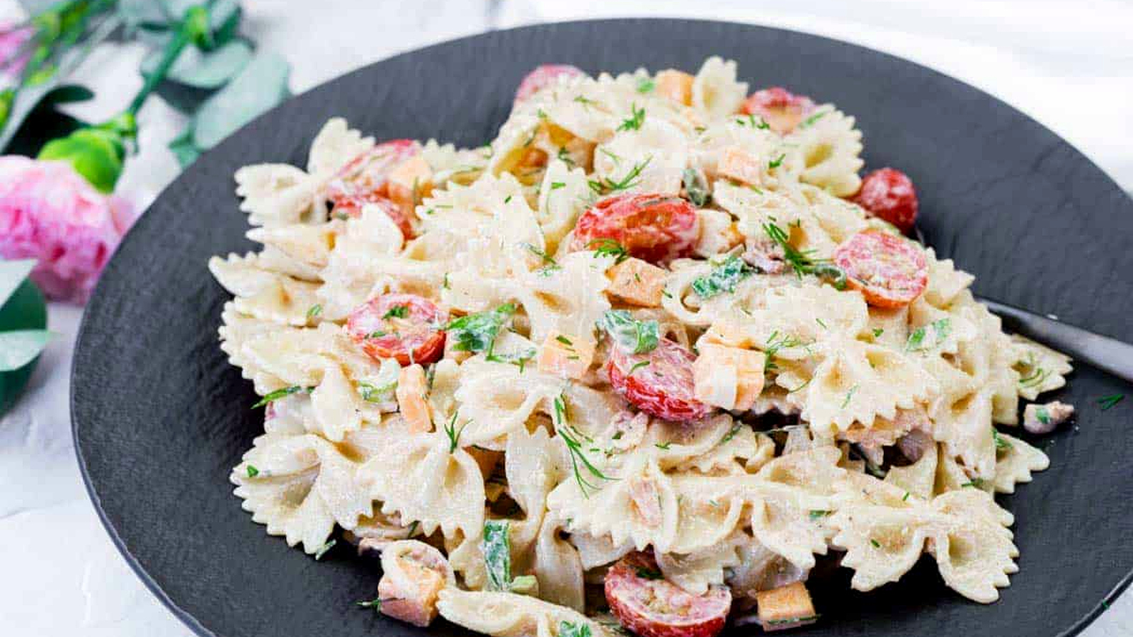 A black plate with bow-tie pasta salad mixed with cherry tomatoes, diced vegetables, herbs, and a creamy dressing.