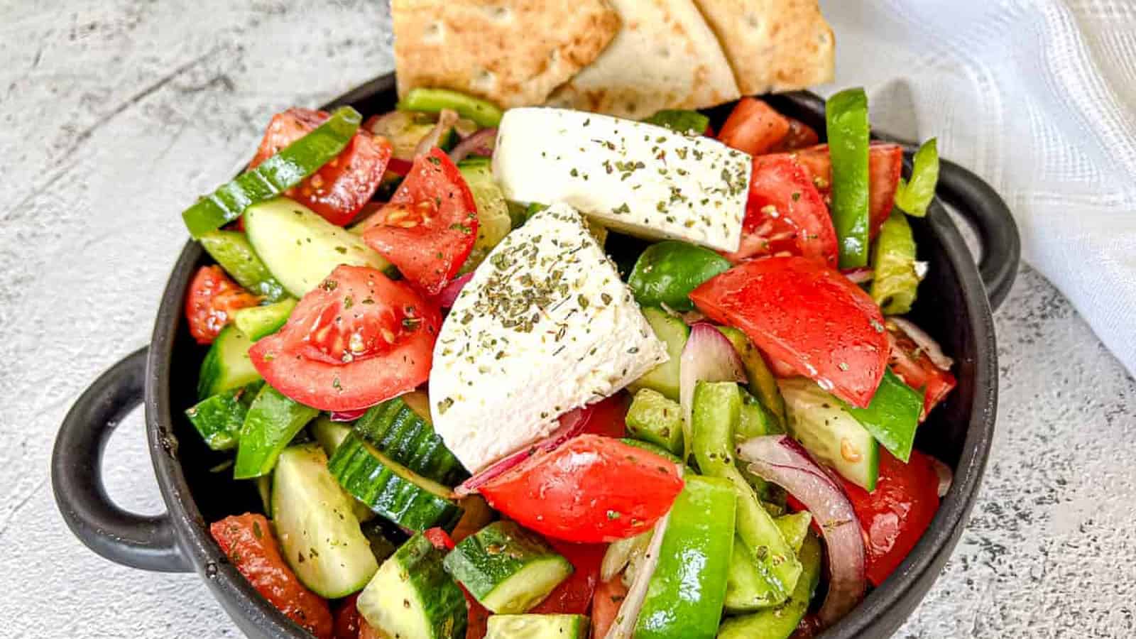 A black bowl contains a Horiatiki salad with blocks of feta cheese, garnished with herbs.