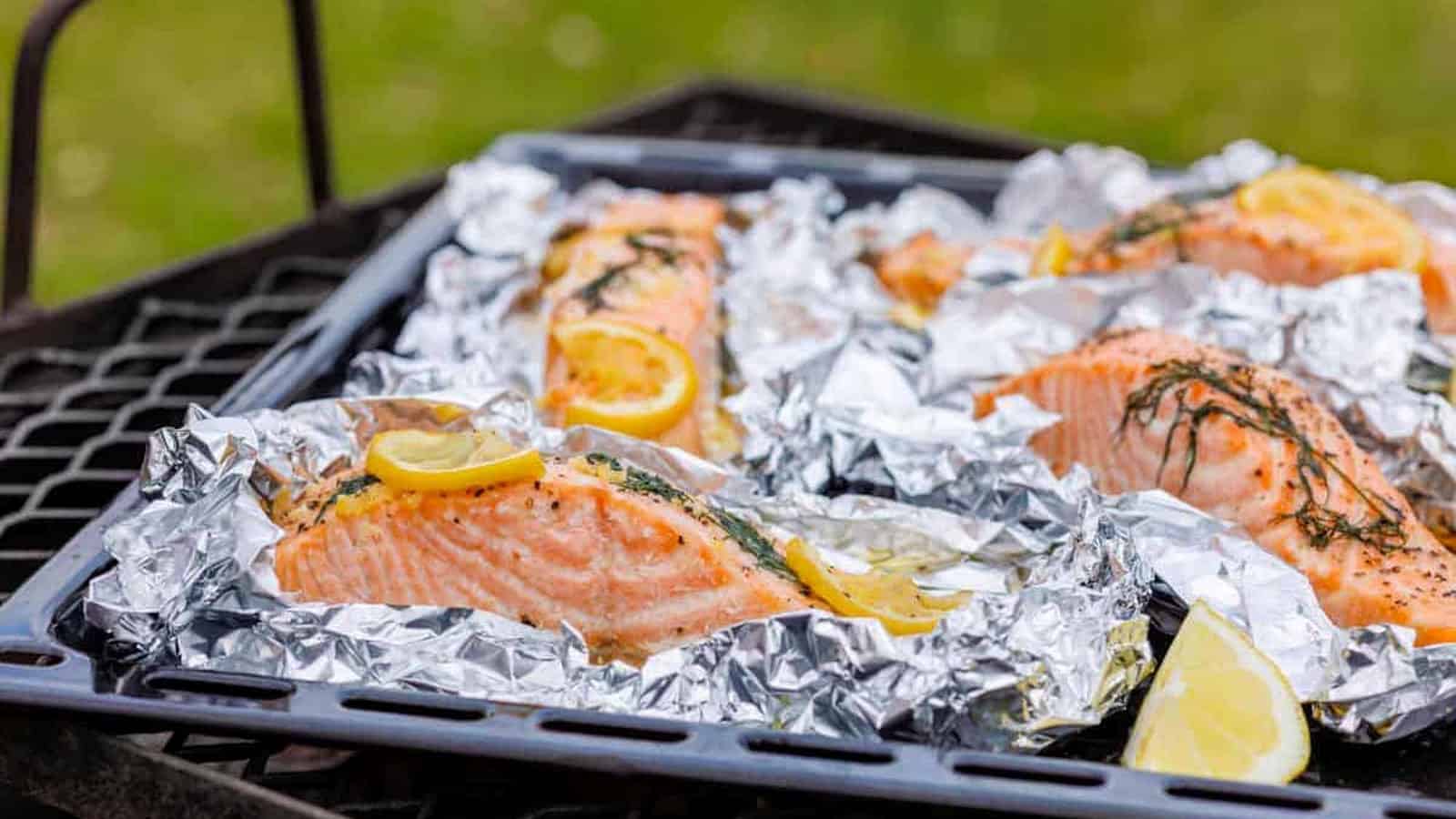 Four servings of lemon-dill salmon in foil packets, with lemon slices and dill on top, arranged on a black grill grate with a green grass background.