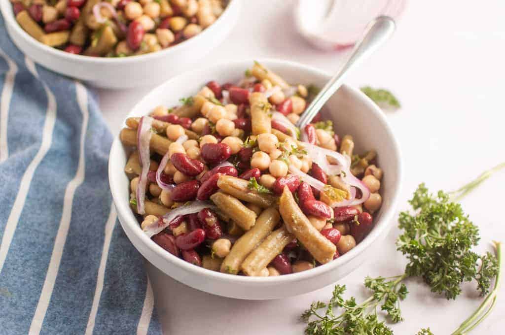 A white bowl filled with a bean salad containing chickpeas, kidney beans, green beans, sliced red onions, and herbs, with a fork inside the bowl.