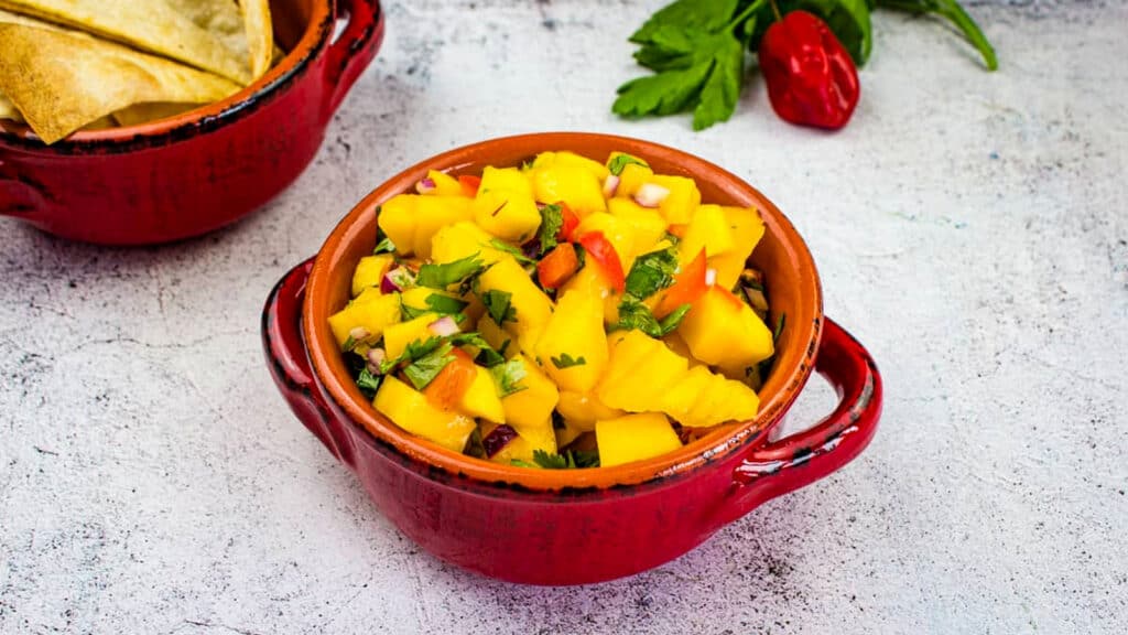 A red bowl filled with mango salsa containing diced mango, red onion, tomato, and cilantro, with tortilla chips and herbs in the background.