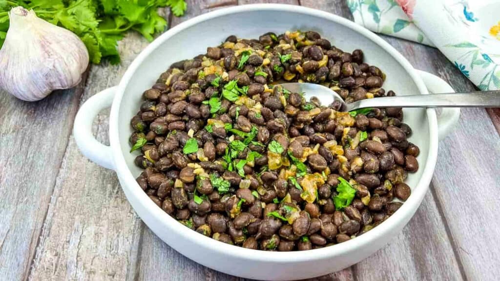 A white bowl filled with cooked black beans garnished with chopped herbs, with a spoon resting inside the bowl. Garlic, cilantro, and a napkin are nearby on a wooden surface.