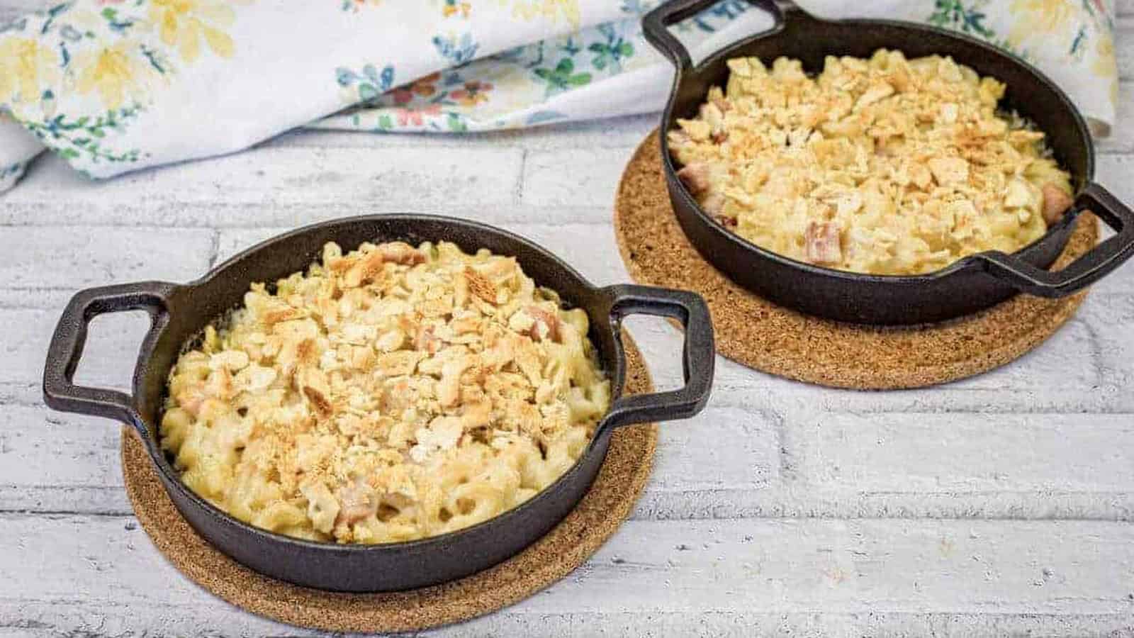Two black cast iron pans filled with mac and cheese sit on cork trivets on a white wooden surface.