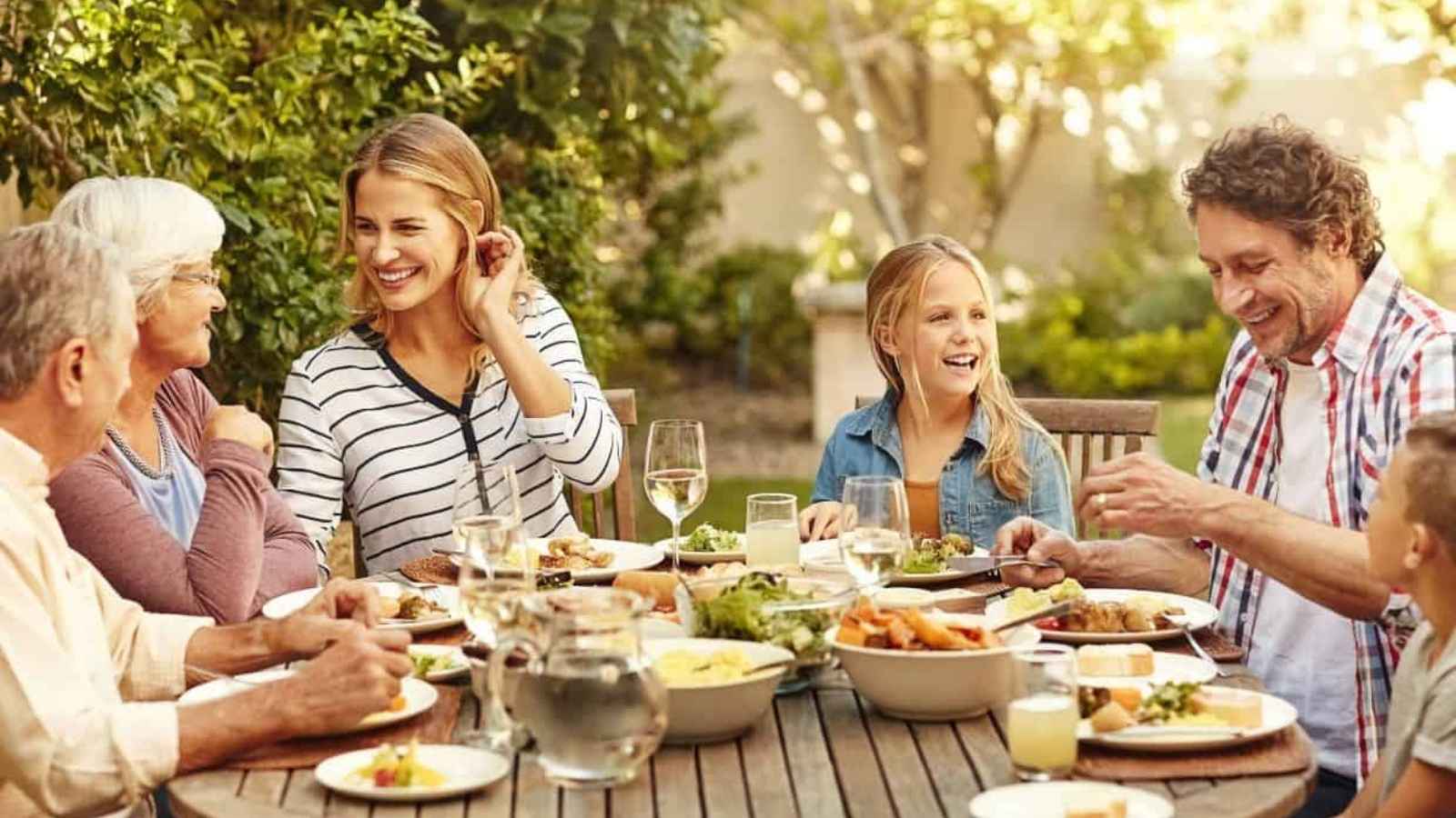 A group of people of various ages sit around an outdoor table, smiling and talking while sharing a meal in a garden setting.