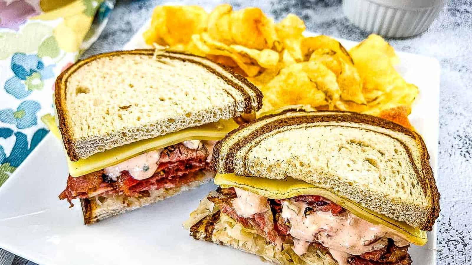 A Pastrami Reuben sandwich on a plate with chips and pickles.