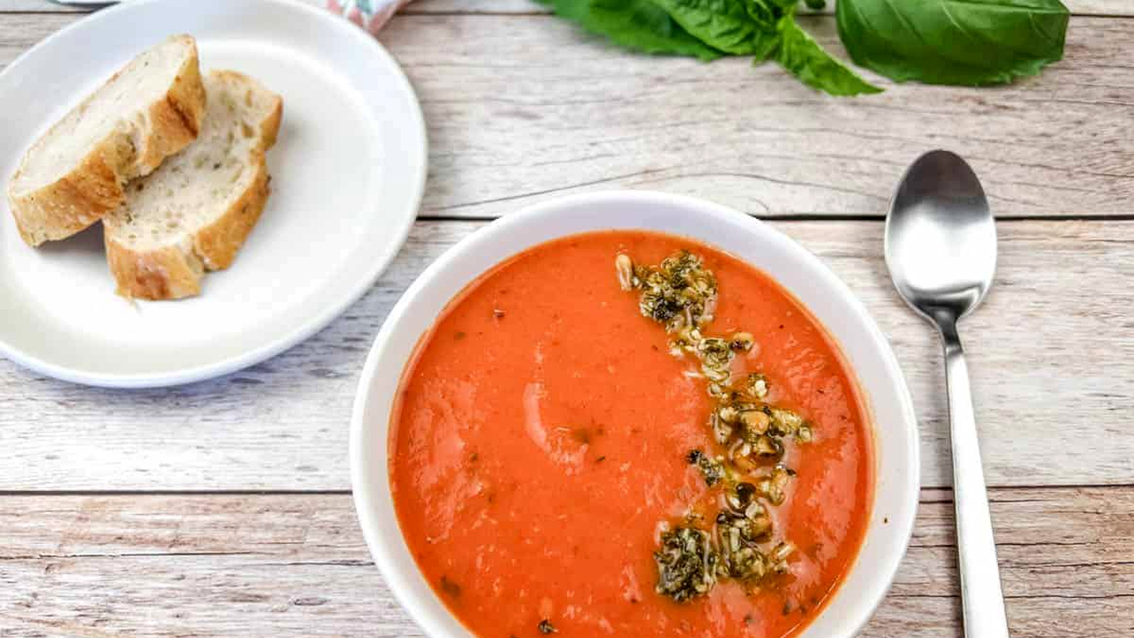 A bowl of red pepper and tomato soup next to a plate of bread and a spoon.