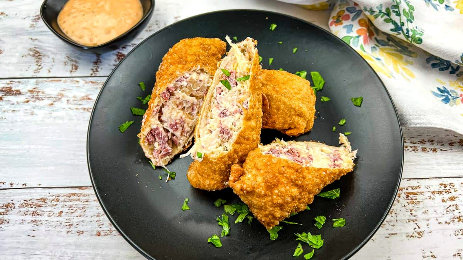 A black plate with Reuben Egg Rolls and dipping sauce.