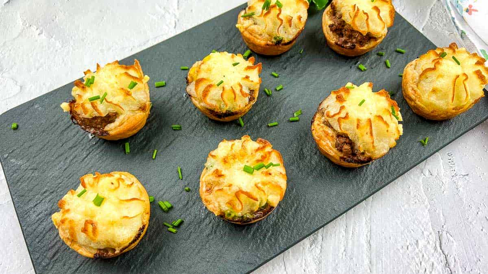 Eight Shepherd's Pie Bites are arranged on a slate board, garnished with chopped chives.