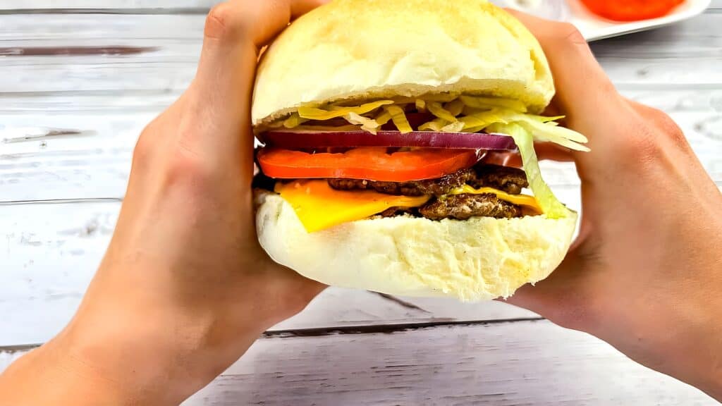 A person holds a hamburger with lettuce, tomato, onion, shredded cheese, cheddar cheese, and a beef patty in a bun over a white wooden surface.