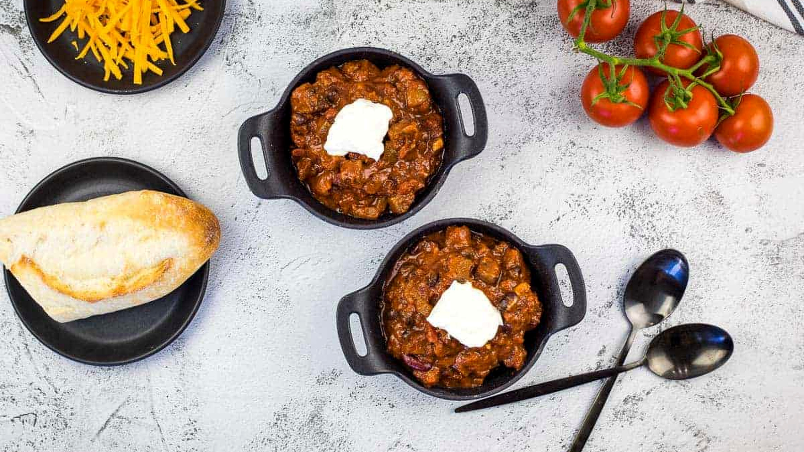 Smoked brisket chili served in two black bowls topped with sour cream, surrounded by fresh tomatoes, shredded cheese, crusty bread, and two spoons on a gray surface.