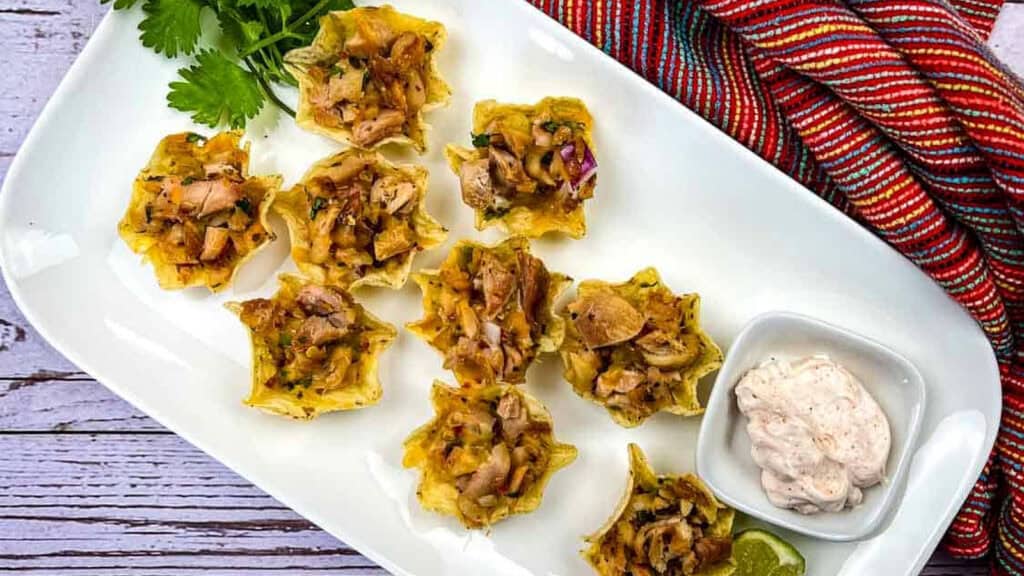 A white plate with eight chicken-filled tortilla cups, a small dish of creamy dip, a lime wedge, cilantro garnish, and a colorful striped cloth on the side.