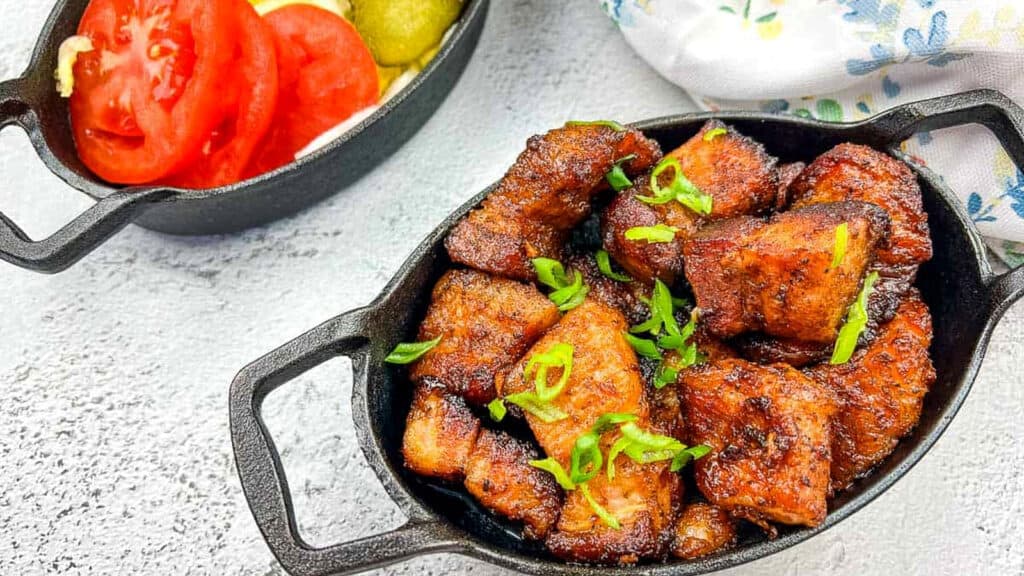 Cast iron dish with browned, seasoned chicken pieces garnished with chopped green onions; sliced tomatoes, onions, and pickles in a separate small dish nearby.