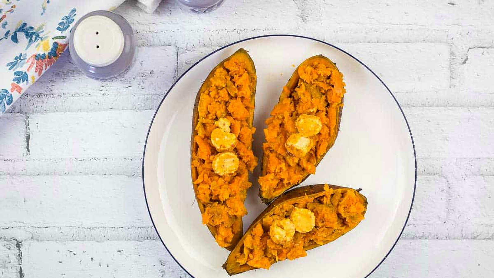 Three halves of roasted sweet potatoes topped with butter on a white plate, set on a white brick surface with a floral napkin and salt and pepper shakers nearby.
