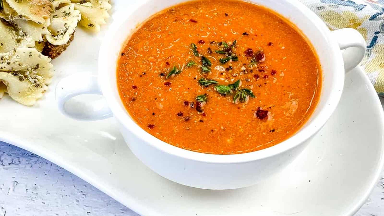 A bowl of smoked tomato soup with mascarpone, garnished with herbs and chili flakes, served with seasoned pasta on a white plate.