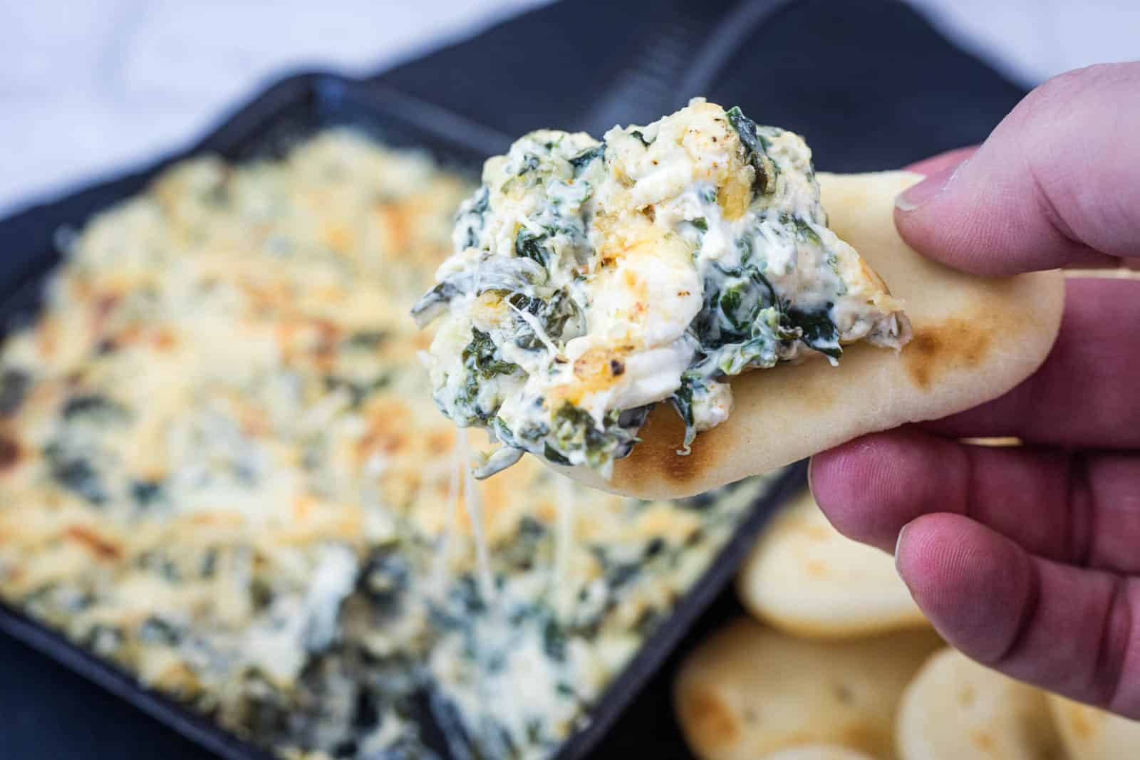 A hand holds a piece of flatbread topped with creamy spinach artichoke dip. A baking dish with more dip and a stack of flatbreads are in the background.