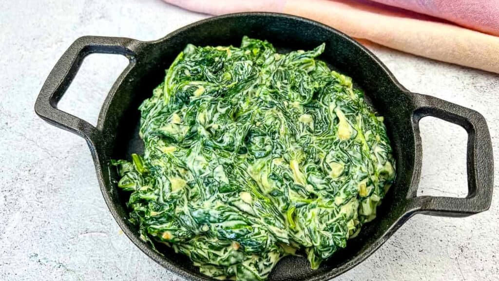 A small cast iron skillet filled with creamy creamed spinach sits on a light surface with a pink cloth in the background.
