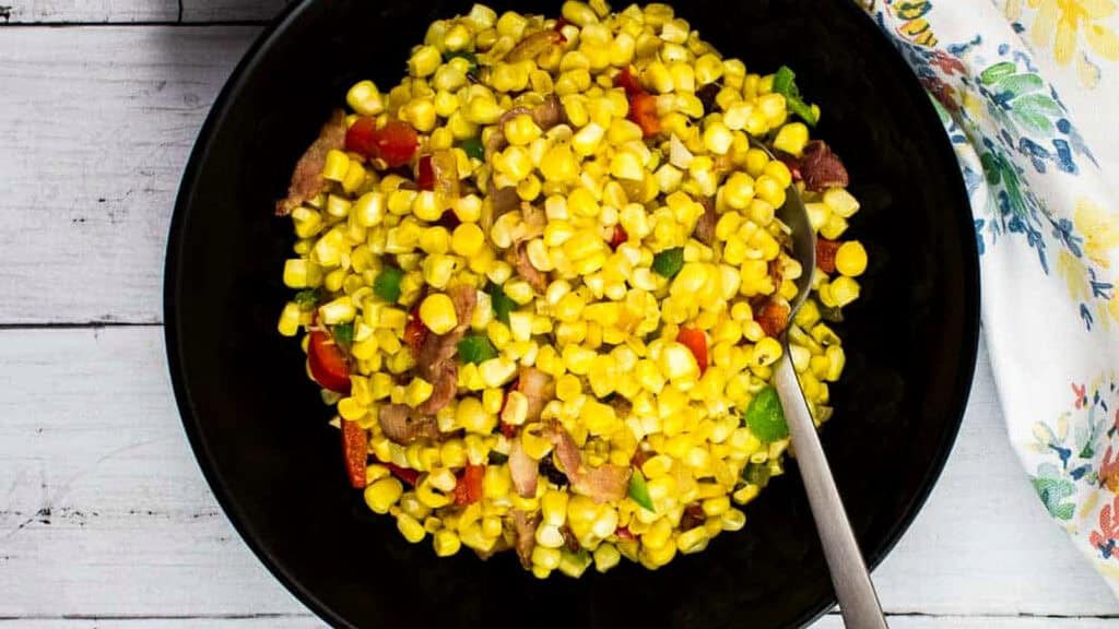 A black bowl filled with corn salad containing corn kernels, diced vegetables, and bacon pieces, with a metal spoon resting inside.