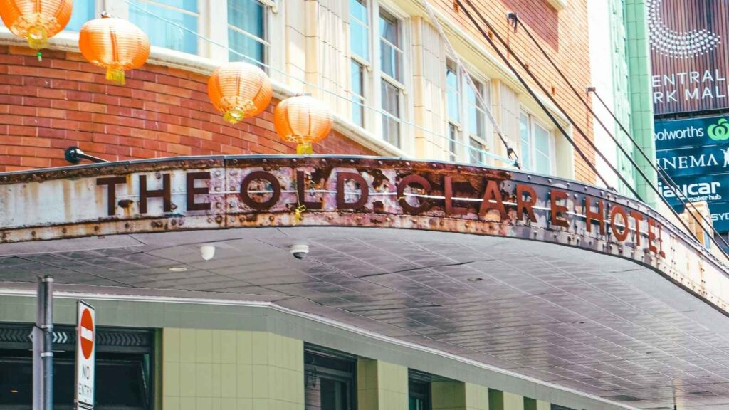 Rusty marquee sign reading "The Old Clare Hotel" with orange lanterns hanging above, attached to a brick building on a city street.