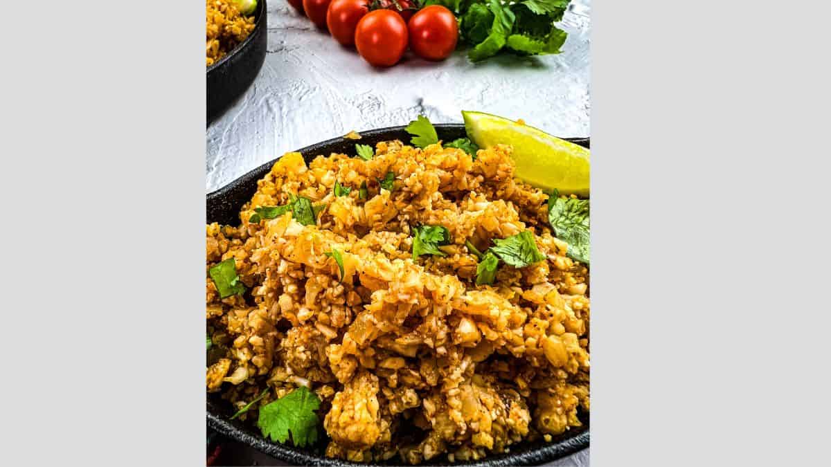 A plate of Mexican Cauliflower Rice garnished with cilantro, served with a lime wedge. Fresh tomatoes and cilantro are visible in the background.