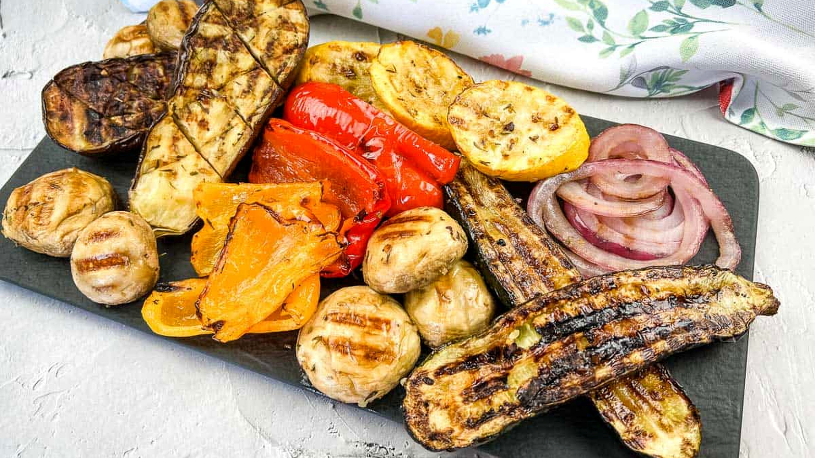 Grilled vegetables including zucchini, mushrooms, red onions, peppers, and squash, arranged on a rectangular black platter.