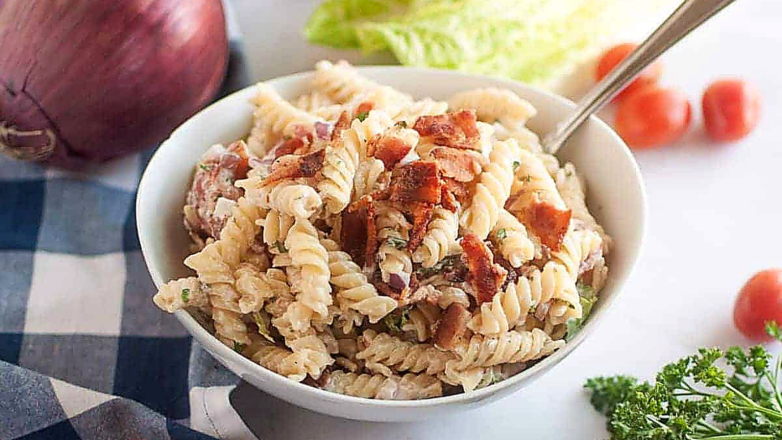 A white bowl filled with BLT pasta salad, featuring spiral pasta, creamy dressing, pieces of bacon, and other visible ingredients, is shown with a spoon. Red onions, lettuce, cherry tomatoes, and parsley are in the background.