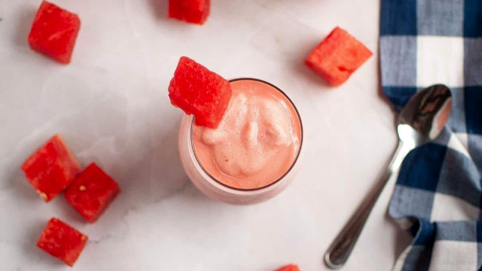 A glass of watermelon smoothie topped with a watermelon cube, surrounded by watermelon pieces, a spoon, and a blue-checkered cloth on a white surface.