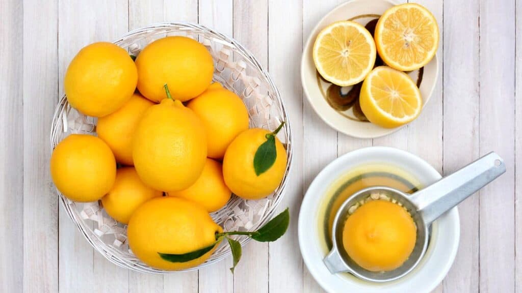 A basket of whole lemons, a plate with halved lemons, and a lemon being juiced on a manual citrus juicer, all on a light wooden surface.
