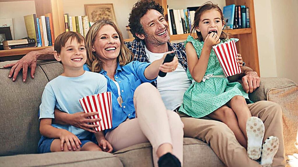 A family of four sits on a couch eating popcorn and watching TV together, smiling and enjoying their time in a cozy living room.