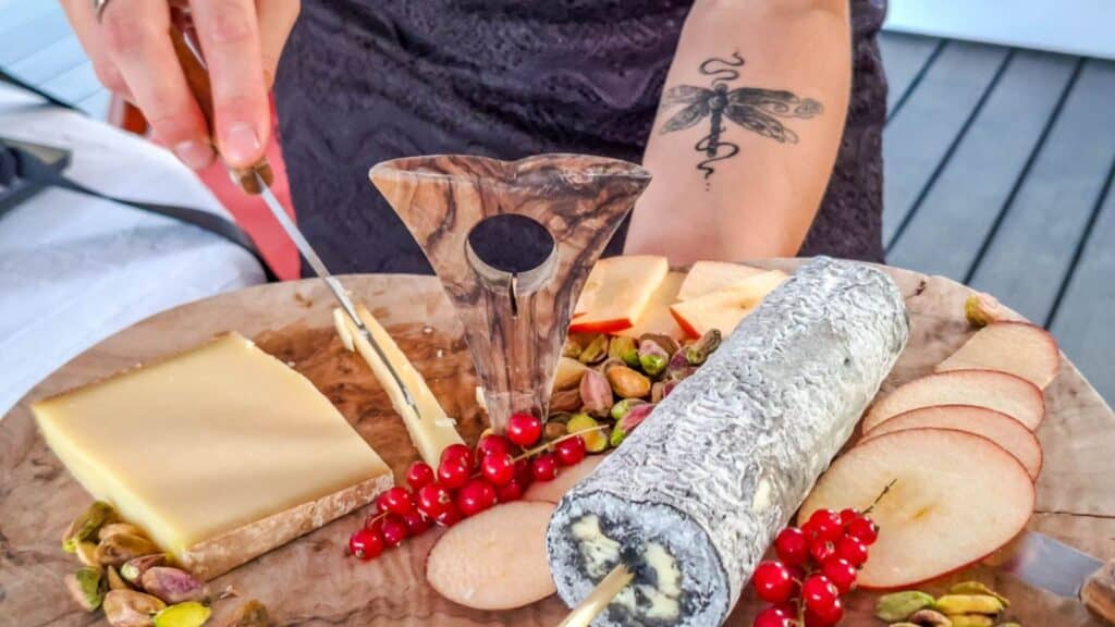 A person slices cheese on a wooden board with assorted cheeses, apple slices, red currants, pistachios, and a cheese knife—evoking the relaxed elegance of a southern France barge cruise. A tattoo is visible on the person’s forearm.