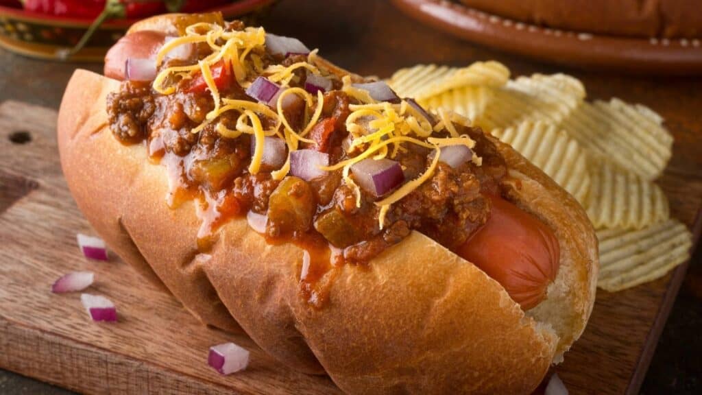 Celebrate National Chili Dog Day with our chili cheese hot dog, topped with shredded cheddar and diced red onions, served alongside ridged potato chips on a wooden board.