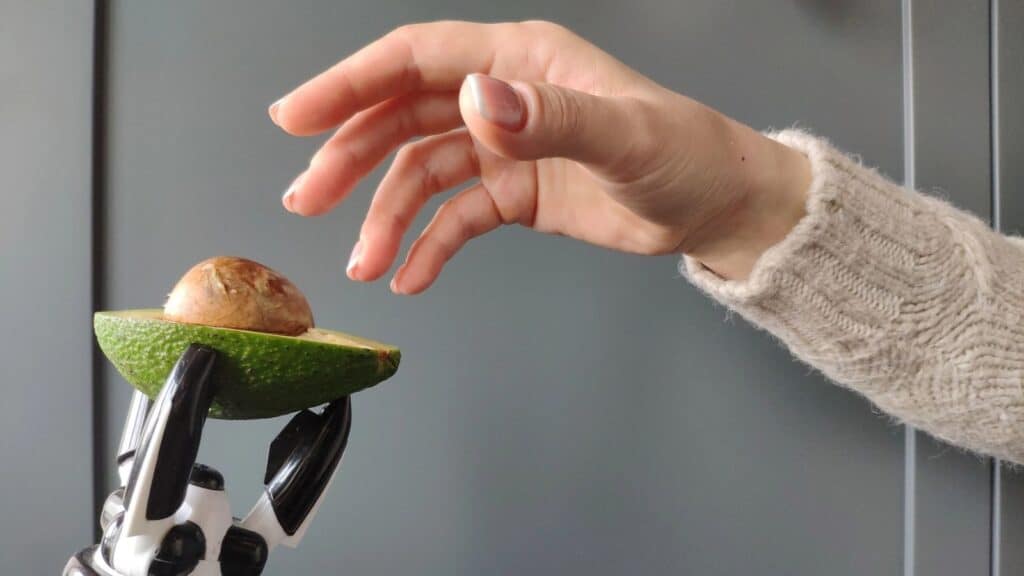 A robotic hand, resembling an avocado-picking robot, holds half an avocado with a pit, while a human hand reaches toward it against a plain background.