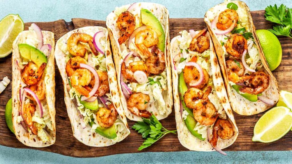 Five shrimp tacos with lettuce, sliced avocado, red onion, and lime wedges are arranged on a wooden board—a perfect pairing for your favorite margarita flavors.