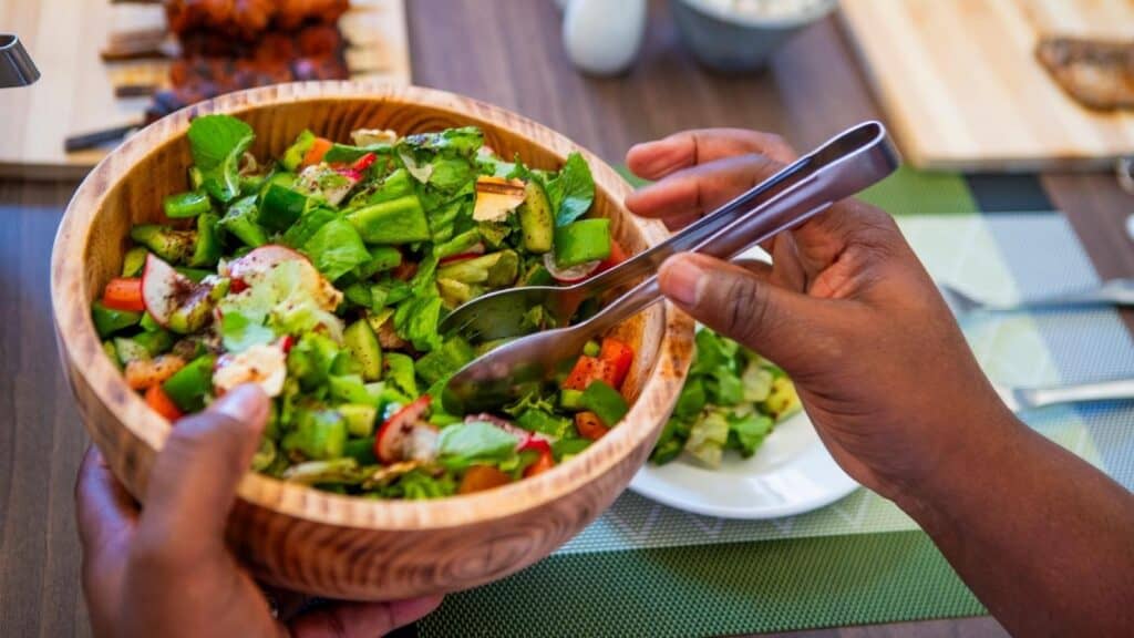 Person in Nairobi holding a wooden bowl of fresh salad with serving tongs, featuring lettuce, tomatoes, cucumbers, and other vegetables on a dining table.