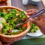 Person in Nairobi holding a wooden bowl of fresh salad with serving tongs, featuring lettuce, tomatoes, cucumbers, and other vegetables on a dining table.