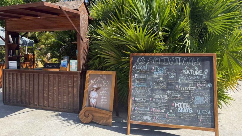 Wooden concierge kiosk beside a large chalkboard listing daily activities in Spanish, with palm plants in the background and sunlight casting shadows&mdash;an inviting scene often found at family wellness resorts.