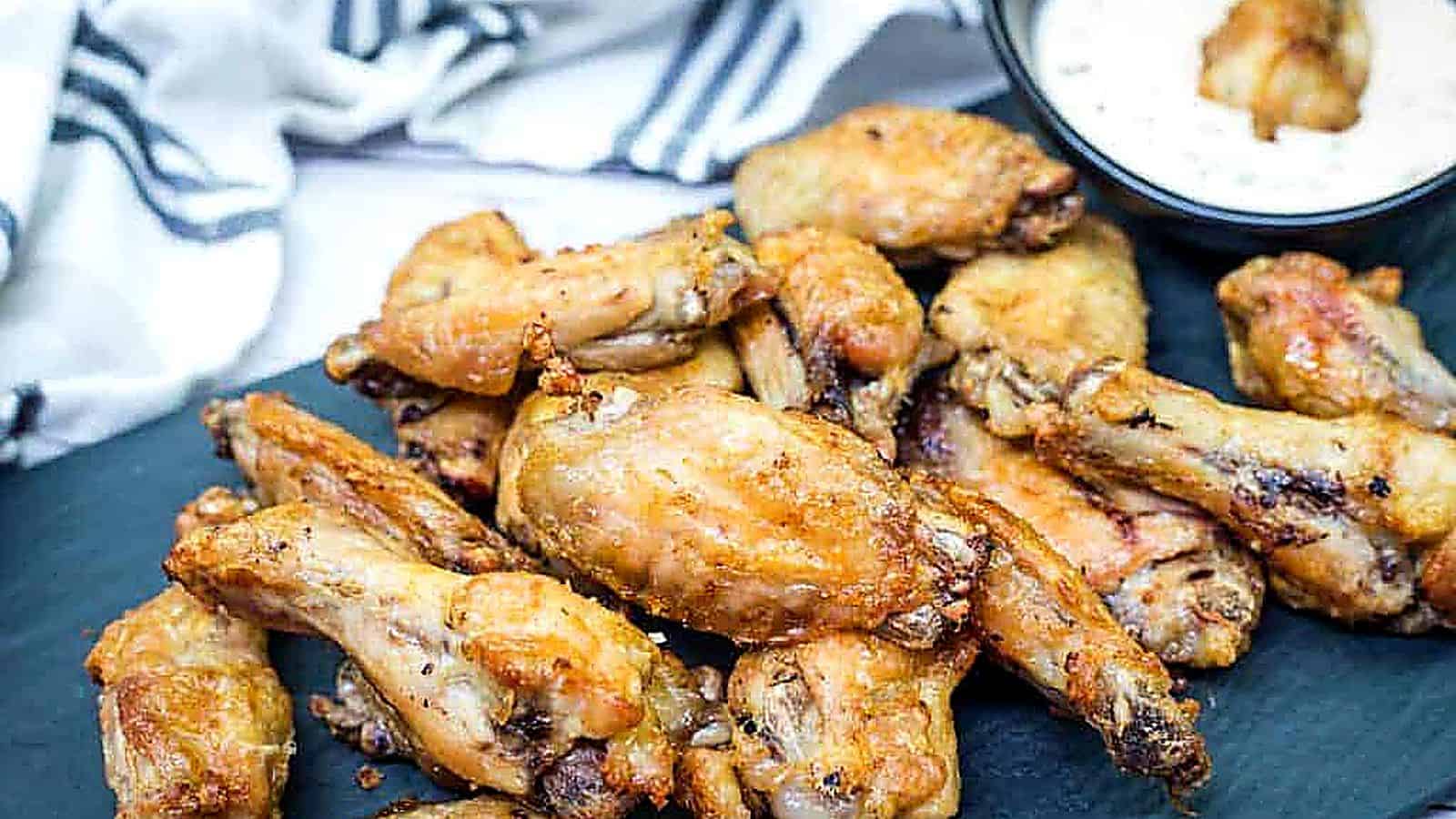 A pile of air-fried chicken wings on a black slate platter with a bowl of creamy dipping sauce in the background.