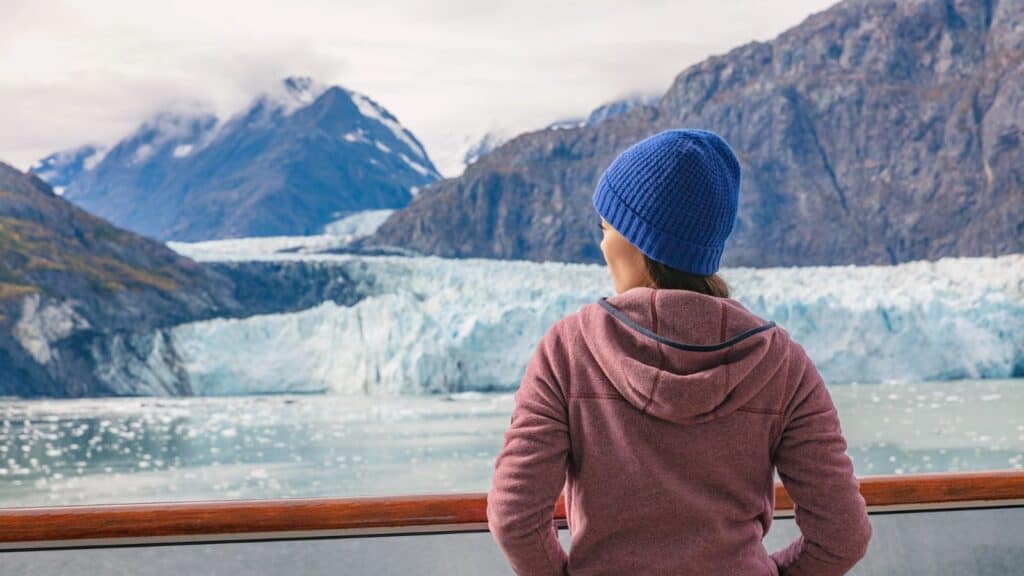 Person in a blue beanie and pink hoodie stands on a deck, looking at a glacier and snow-capped mountains across a body of water.