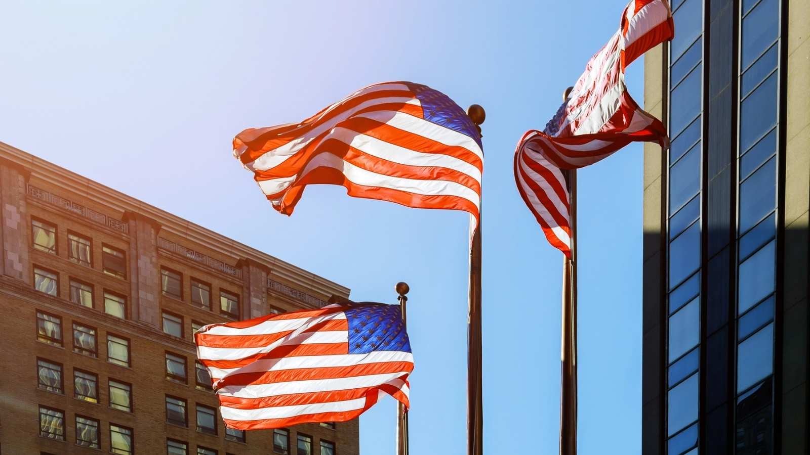 Three American flags wave in the wind, celebrating the nation&rsquo;s 250th anniversary, in front of modern and historic buildings under a clear, sunny sky.