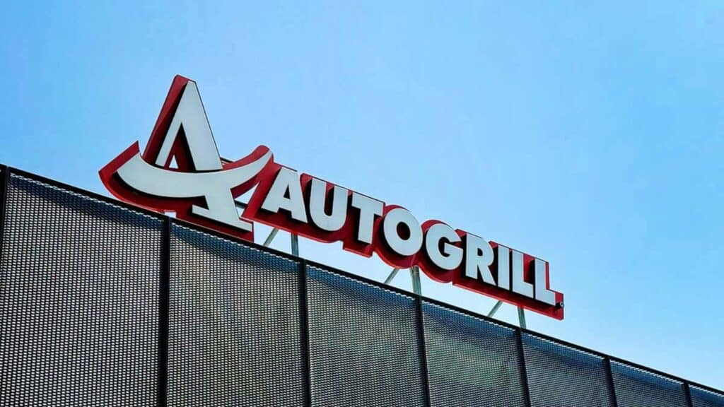 A large red and white autogrill sign is mounted on top of a perforated metal structure, set against a clear blue sky.