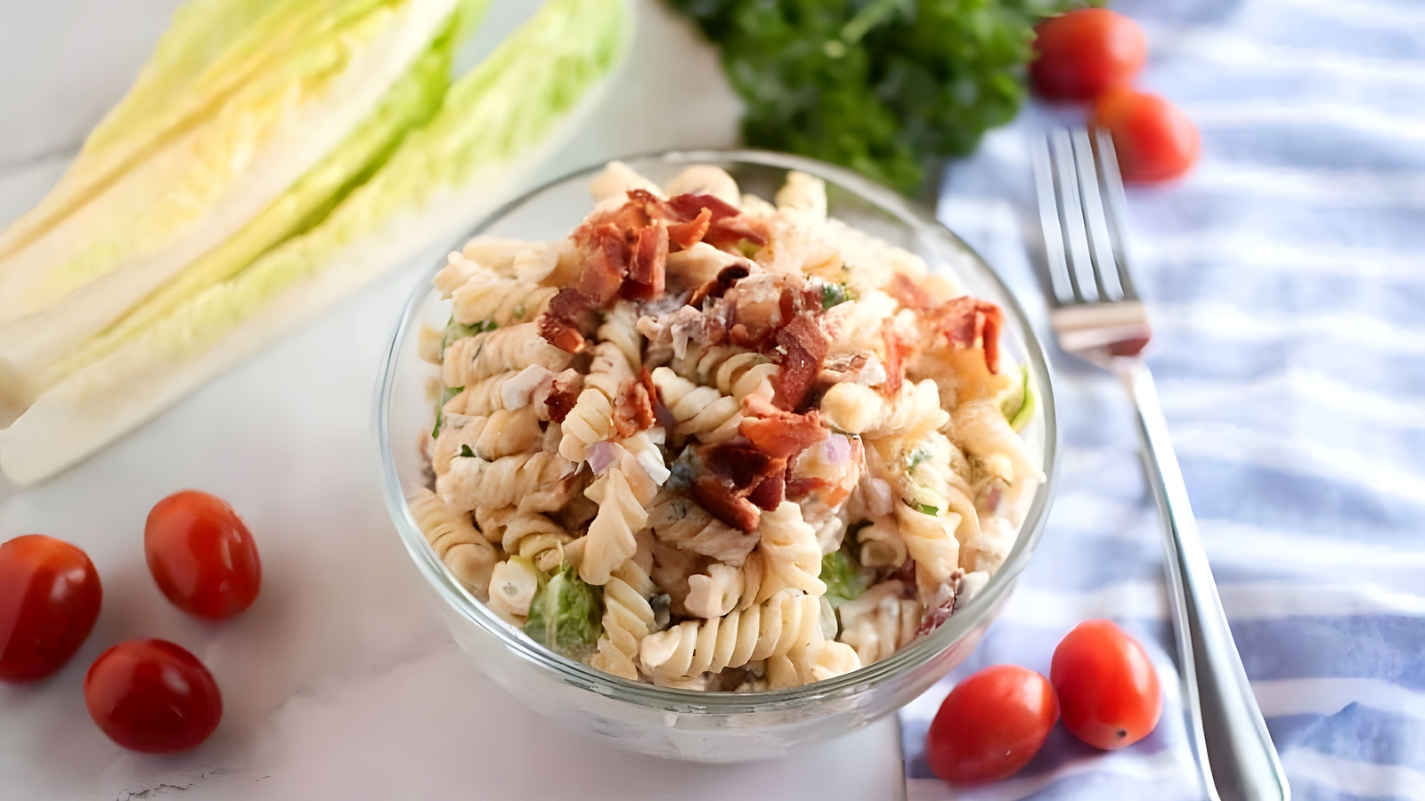 A glass bowl of rotini pasta salad topped with bacon, surrounded by grape tomatoes, lettuce, a fork, and parsley on a striped cloth.