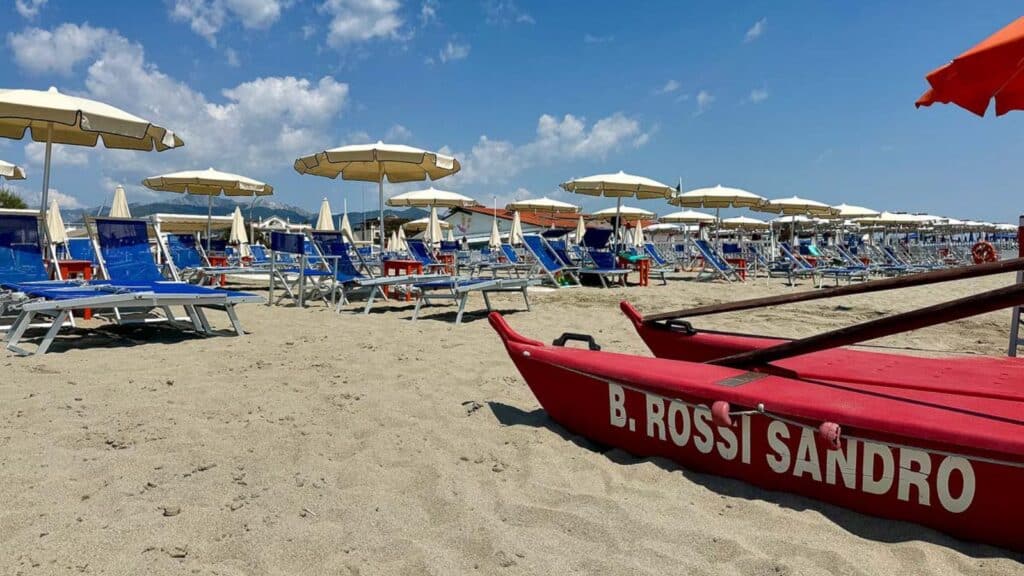 Rows of blue lounge chairs and beach umbrellas line the sandy beach, with a red lifeboat labeled "B. Rossi Sandro" in the foreground under a partly cloudy sky.