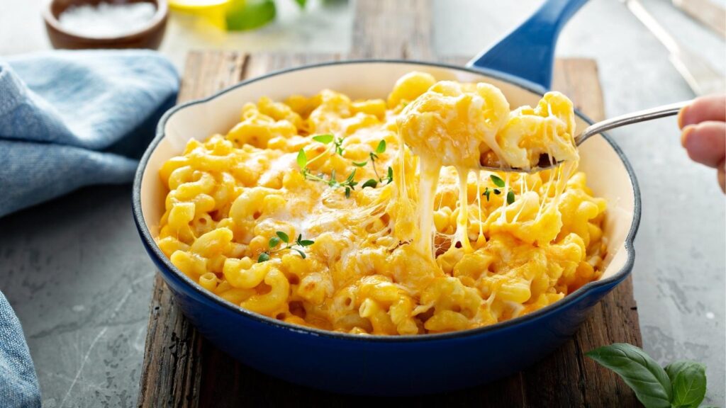 Baked mac and cheese in a blue skillet, topped with melted cheese and herbs, with a spoon lifting a cheesy portion.